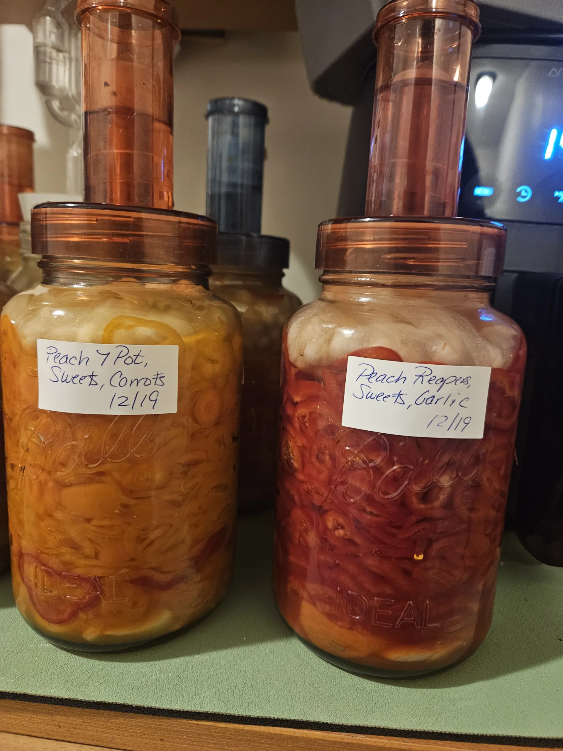 Fermentation jars with airlocks. The jars are filled with sliced peppers. One jar is labeled Peach, 7 Pot, Sweets, Carrots, 12/19. The other jar is labeled Peach Reapers, Sweets, Garlic, 12/19